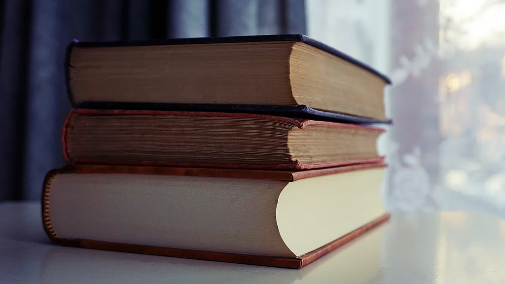 A stack of three old hardcover books on a table by a window